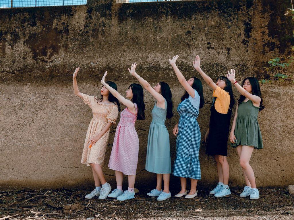 Encompass HK LGBTQ Gay Lesbians Hong Kong HK Mentorship LGBTQ+ Diversity Inclusion Sustainability Sustainable SDGs Sustainable Development Goals SDG Game Responsible Consumption Workshops Belonging D&I DEI Six young women stand in a row against a rough, brown wall, all raising their right hands as if waving or shielding their eyes from sunlight. Their colorful dresses and white sneakers reflect the spirit of inclusion celebrated by Encompass HK and the LGBTQ+ community.